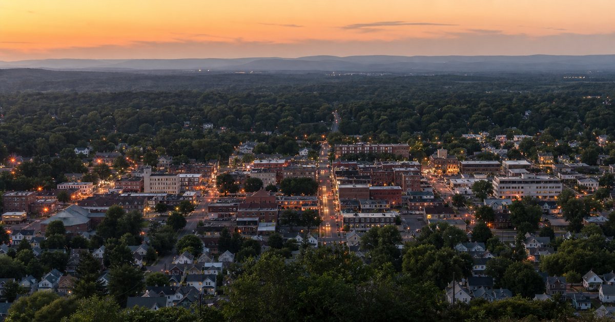 An elevated view of a small American town at dusk seen from a nearby hillside, an orderly grid of tree-lined streets and rooftops with a compact downtown, streetlights beginning to glow, no readable signs